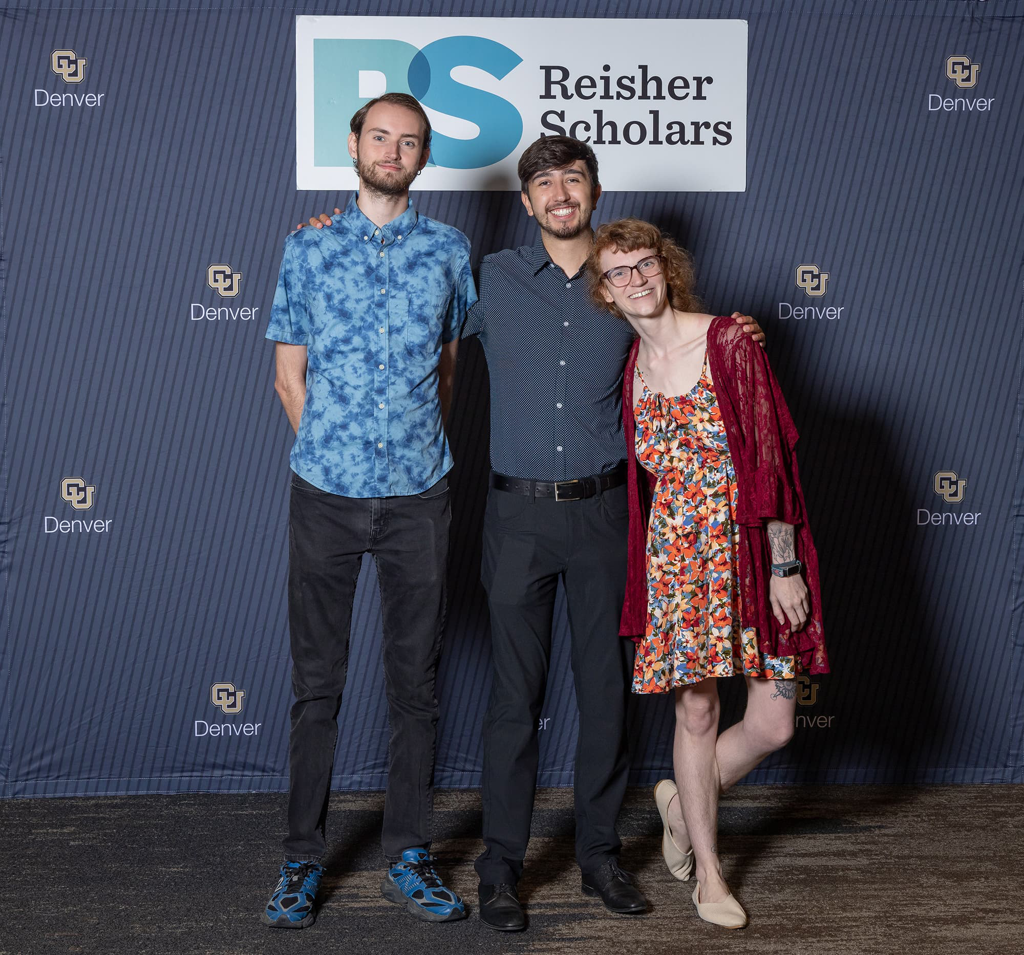 Three students dressed in business attire smile in front of a sign that reads 