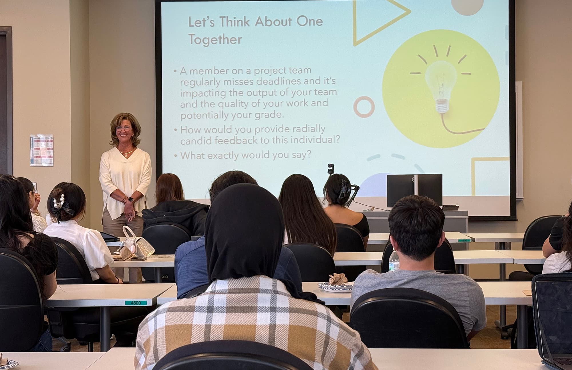 Woman standing in front of a classroom of students with a large PowerPoint screen behind her.