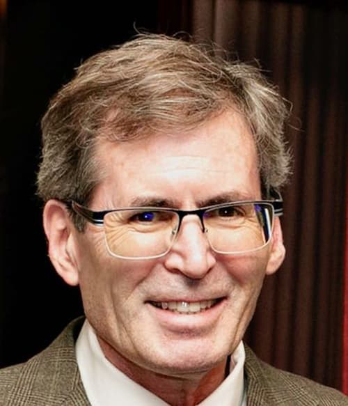 Robert Wolfson in a tan striped suit, square glasses, wavy brown, salt and pepper hair, smiling with his teeth.