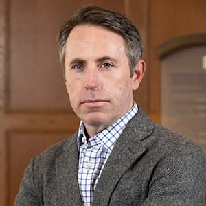 Man in a gray suit jacket and patterned white shirt with a stoic look on his face.