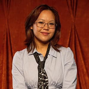 Woman wearing glasses and a closed-mouth smile. She has shoulder-length dark hair and wears a light blouse with a loosely tied tie.