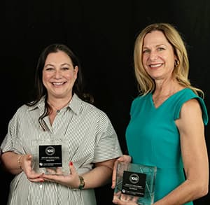 Two women stand against a black background holding plaques and smiling with their teeth.