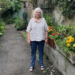 Woman in jeans and a light-colored blouse and shoulder-length white hair stands in front of a garden smiling.