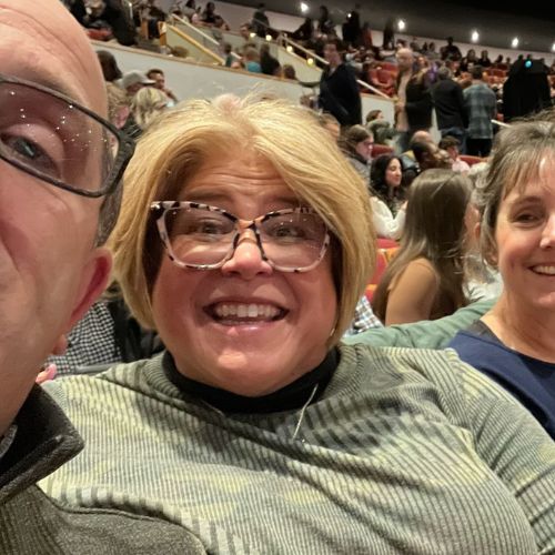 Stacey Huelsing-Dhein smiling broadly while sitting in the audience of a sporting stadium with loved ones.