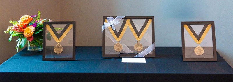 Medals in clear glass frames displayed on a tabletop next to a bouquet of brightly colored flowers before they were presented to awardees.