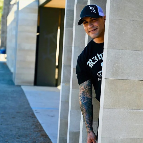 Mario peeks out from behind a concrete wall with a smile on his face. He is wearing a baseball cap and his arm is covered in artistic tattoos.