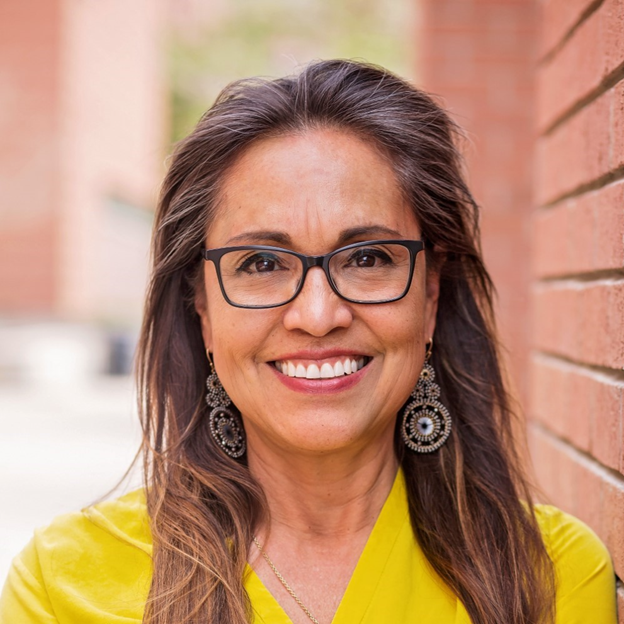 Professional headshot of Theresa Nino; she leans against a brick wall and smiles in a bright yellow top