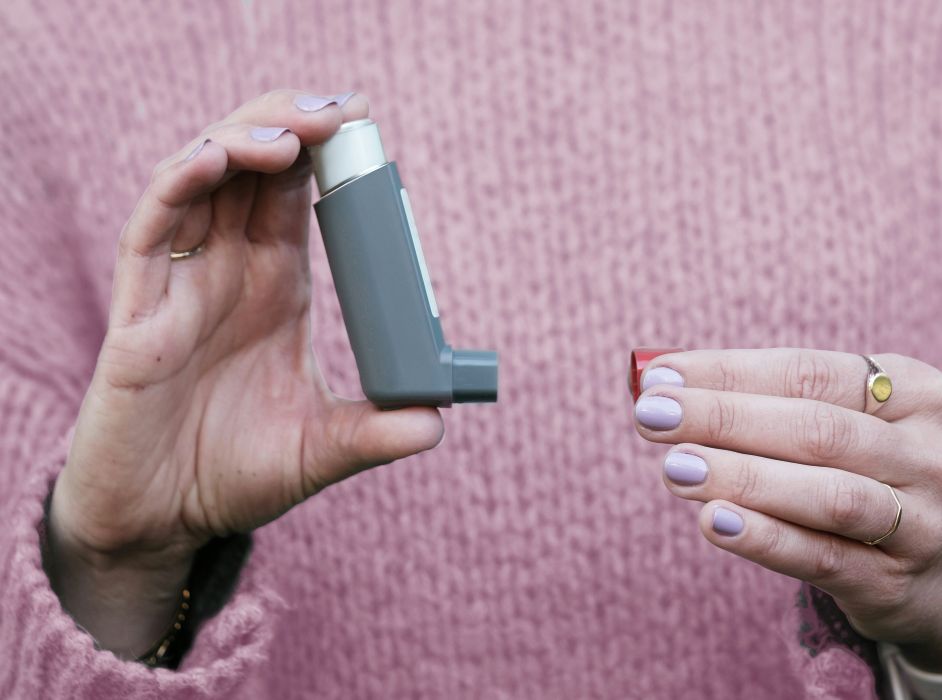 A person with light purple nails holds a gray asthma inhaler in one hand and a red cap in the other, wearing a pink sweater.