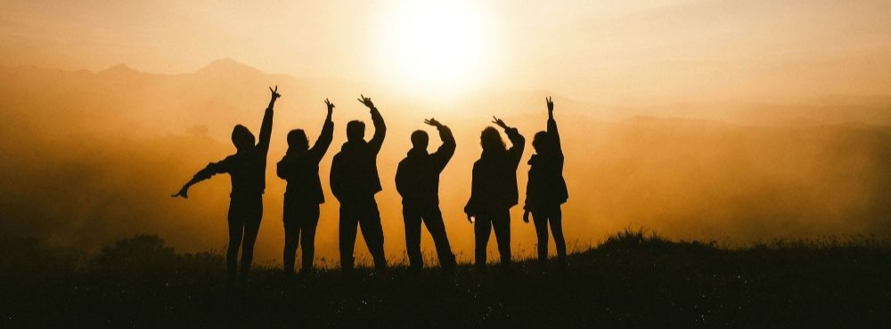 A group of seven people stand in a row on a hill at sunset, silhouetted against the sun with arms raised in various poses.