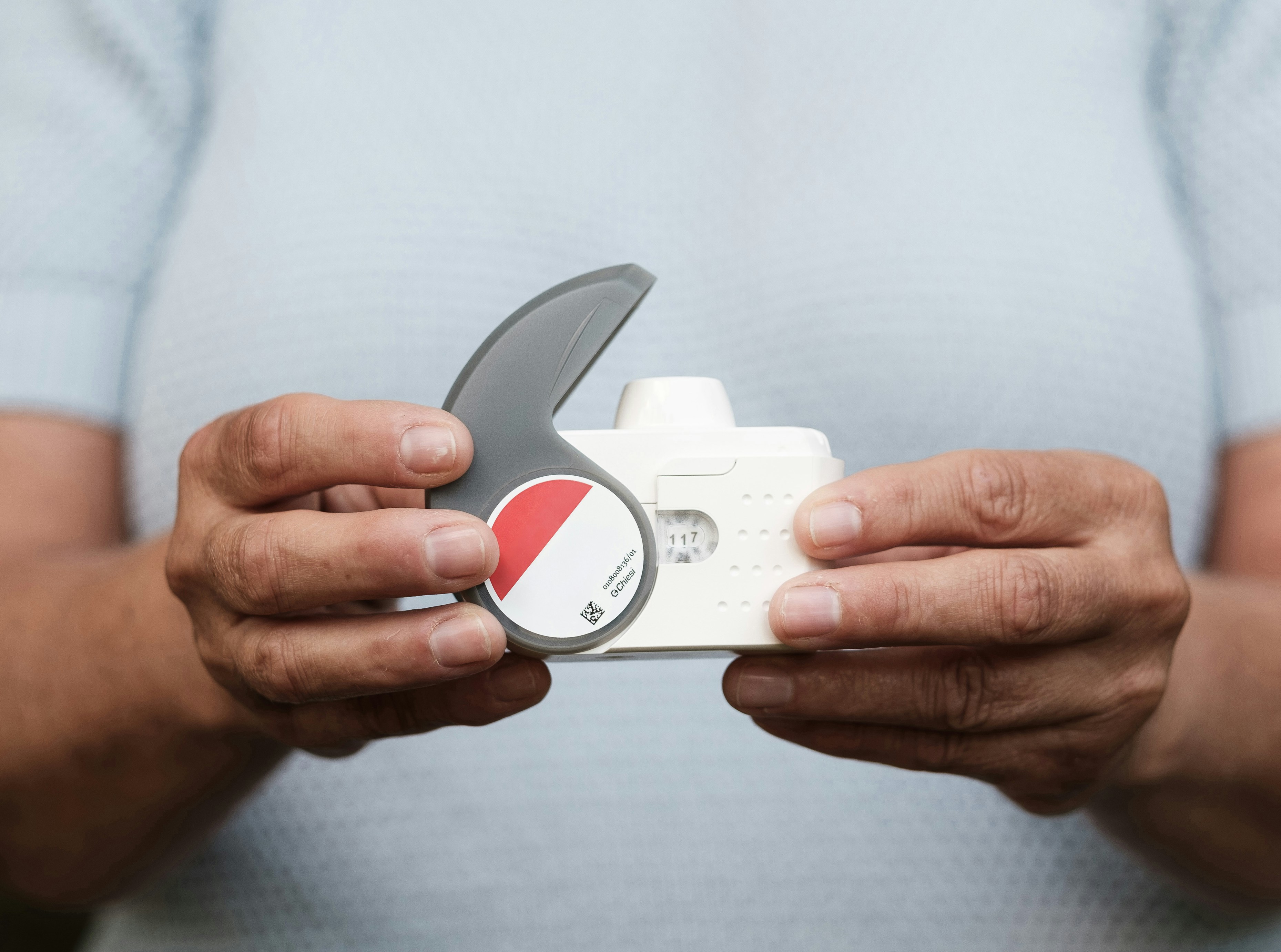 A person holds a white inhaler with a gray handle, featuring a red circular label, against a light blue background.