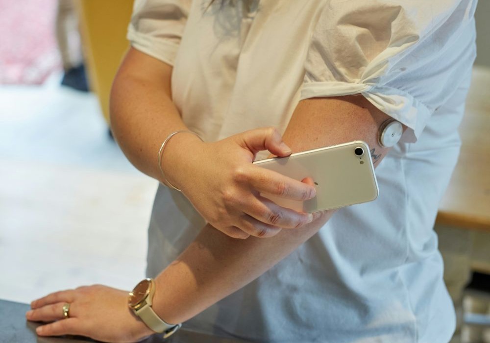 A person checks a continuous glucose monitor attached to their arm with a smartphone, suggesting diabetes management or health monitoring.