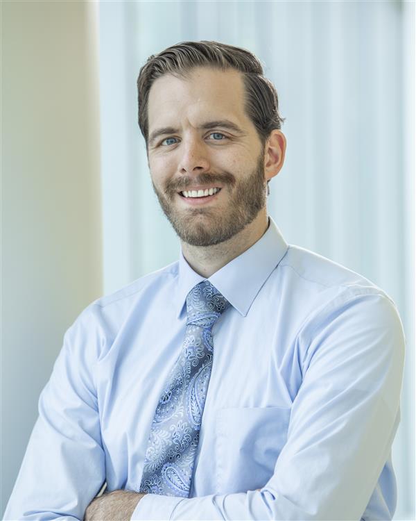 a person with their arms crossed wearing a blue button down with a blue tie with accents, they are smiling for a headshot