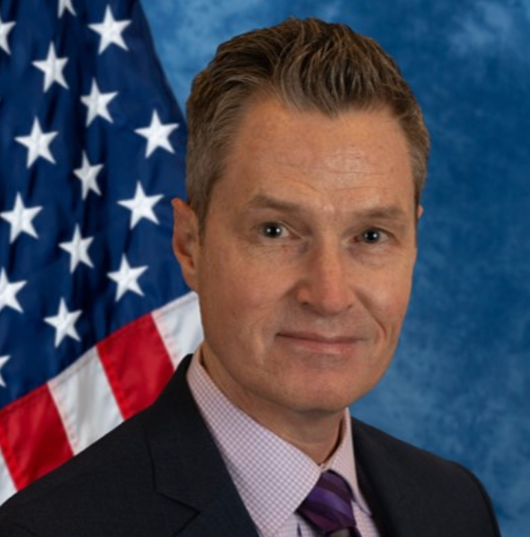 man smiling in a suite in front of a USA flag