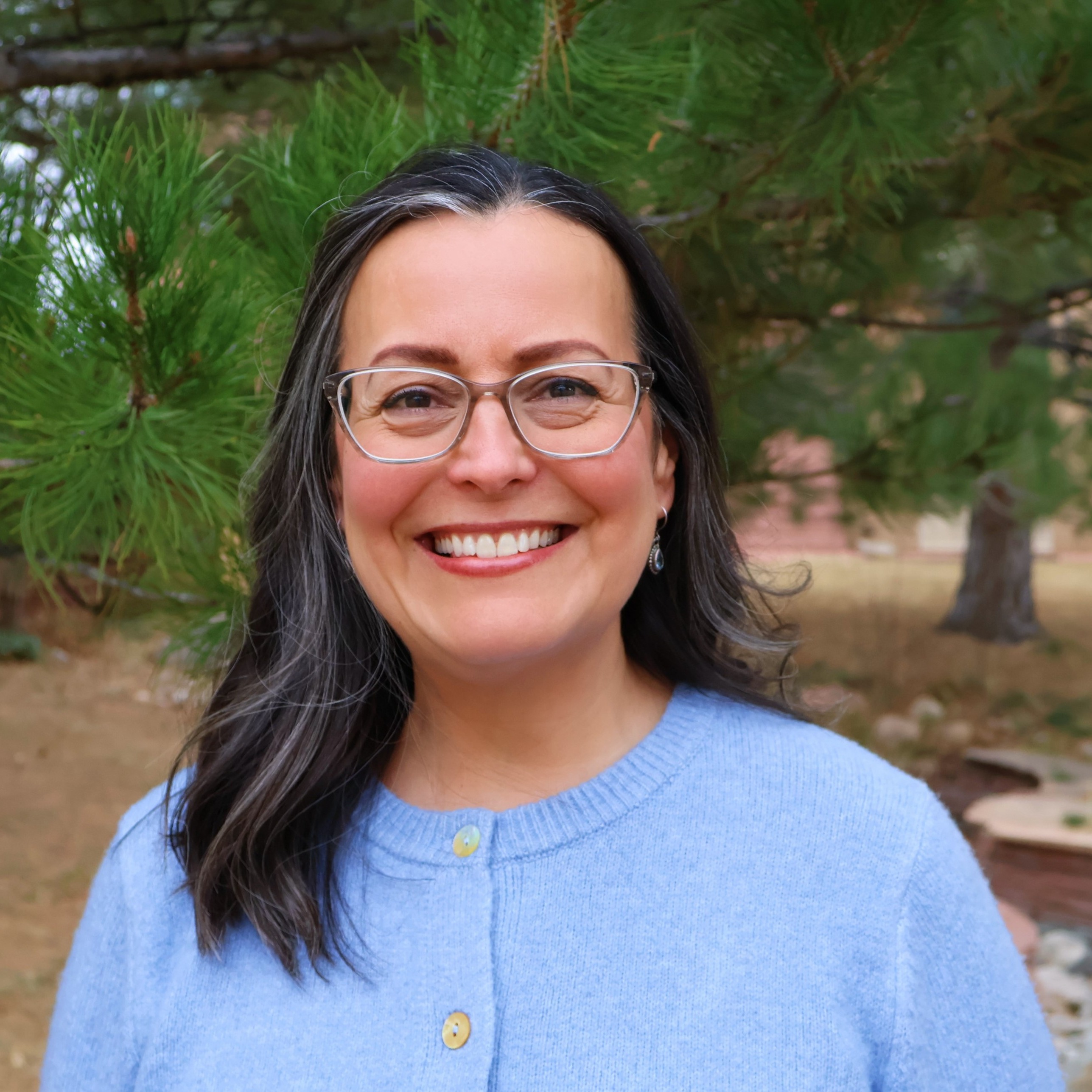 female smiling outside, wearing a light blue button down cardigan, she is wearing glasses and has dark hair