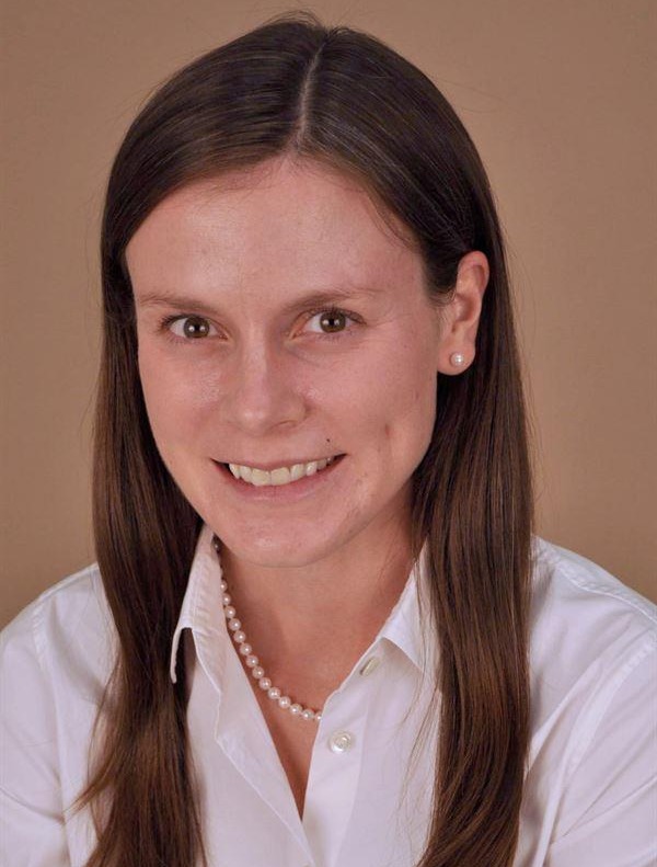 Headshot of a smiling woman with long brown hair wearing a white blouse and pearl necklace against a neutral studio background.