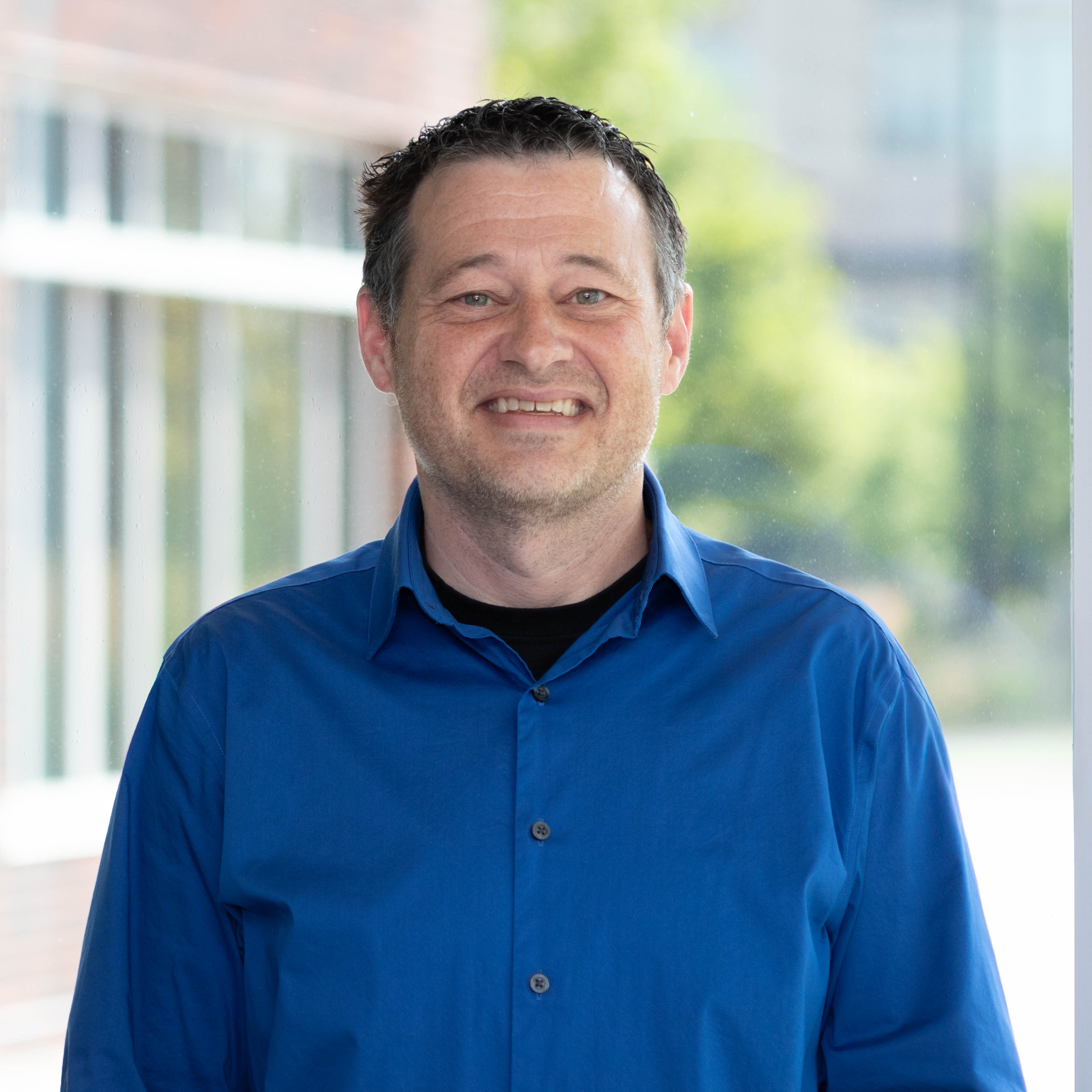 Professional headshot of man in blue collared shirt. Background is a courtyard outside during day, but blurred.