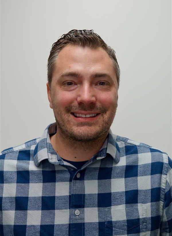 Headshot of a smiling man with short hair and light facial stubble wearing a blue plaid shirt against a plain light background.