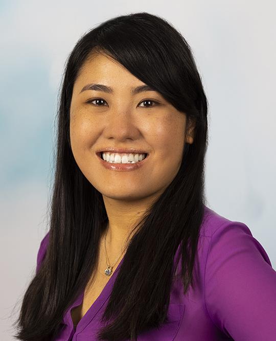 Headshot of a smiling woman with long dark hair wearing a purple blouse and necklace, photographed against a softly blurred background.