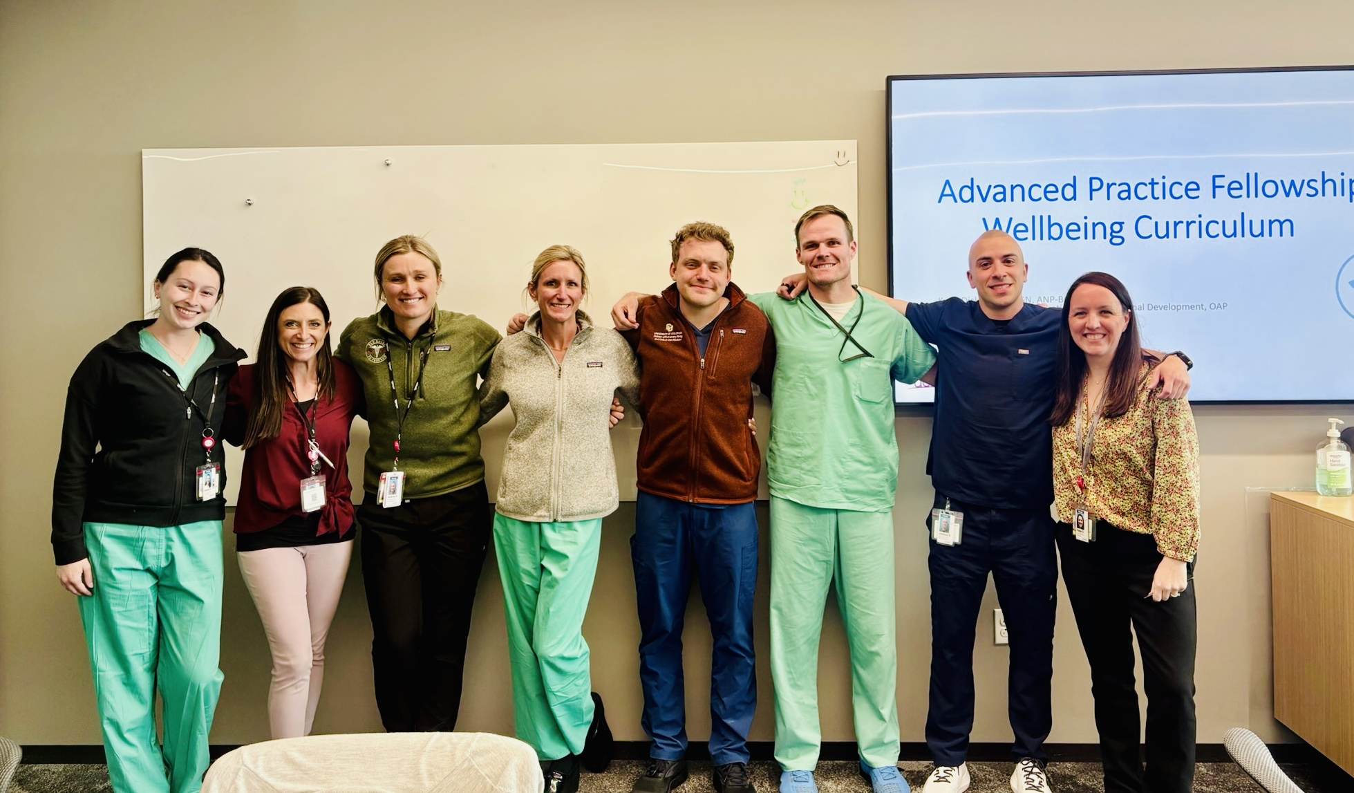 Eight advanced practice providers stand arm in arm in front of a whiteboard and presentation screen displaying “Advanced Practice Fellowship Wellbeing Curriculum” during a fellowship education session.