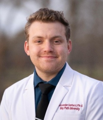 Professional headshot of a smiling person wearing a white medical coat, blue shirt, and black tie, photographed outdoors with a blurred background. The coat includes embroidered name and university affiliation.