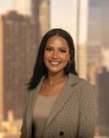 Rachel Machoni.jpg Write alt text  Professional headshot of a smiling woman with long dark hair, wearing a light-colored blazer over a neutral top, standing in front of a blurred city skyline background.