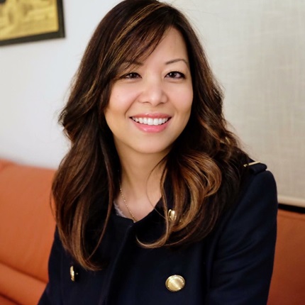 Professional portrait of a smiling person with long wavy hair, wearing a dark blazer with gold buttons, seated indoors against a softly blurred background.