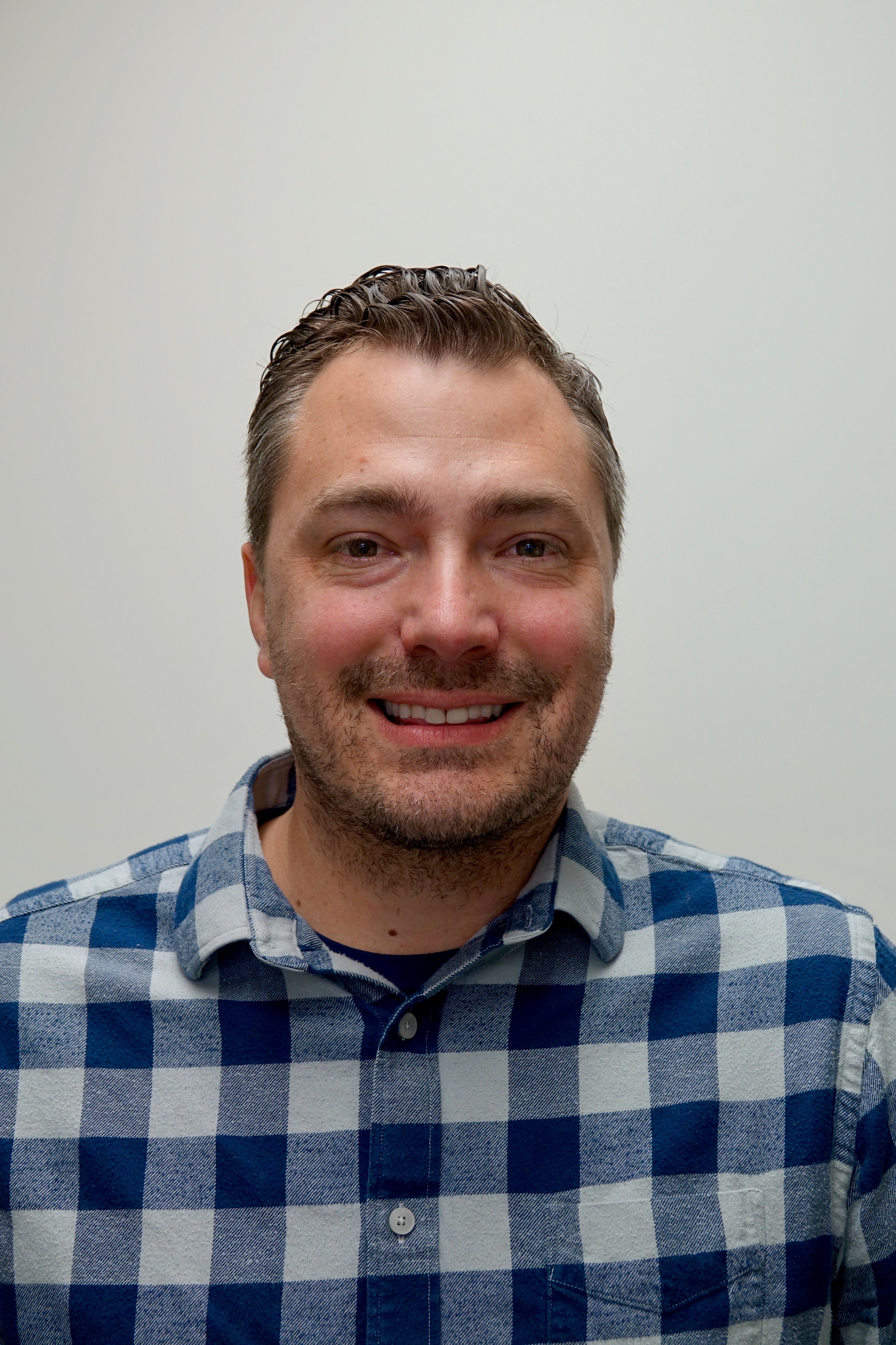 Headshot of a person with short salt-and-pepper hair and light stubble, smiling at the camera while wearing a blue-and-white plaid shirt against a plain light background.