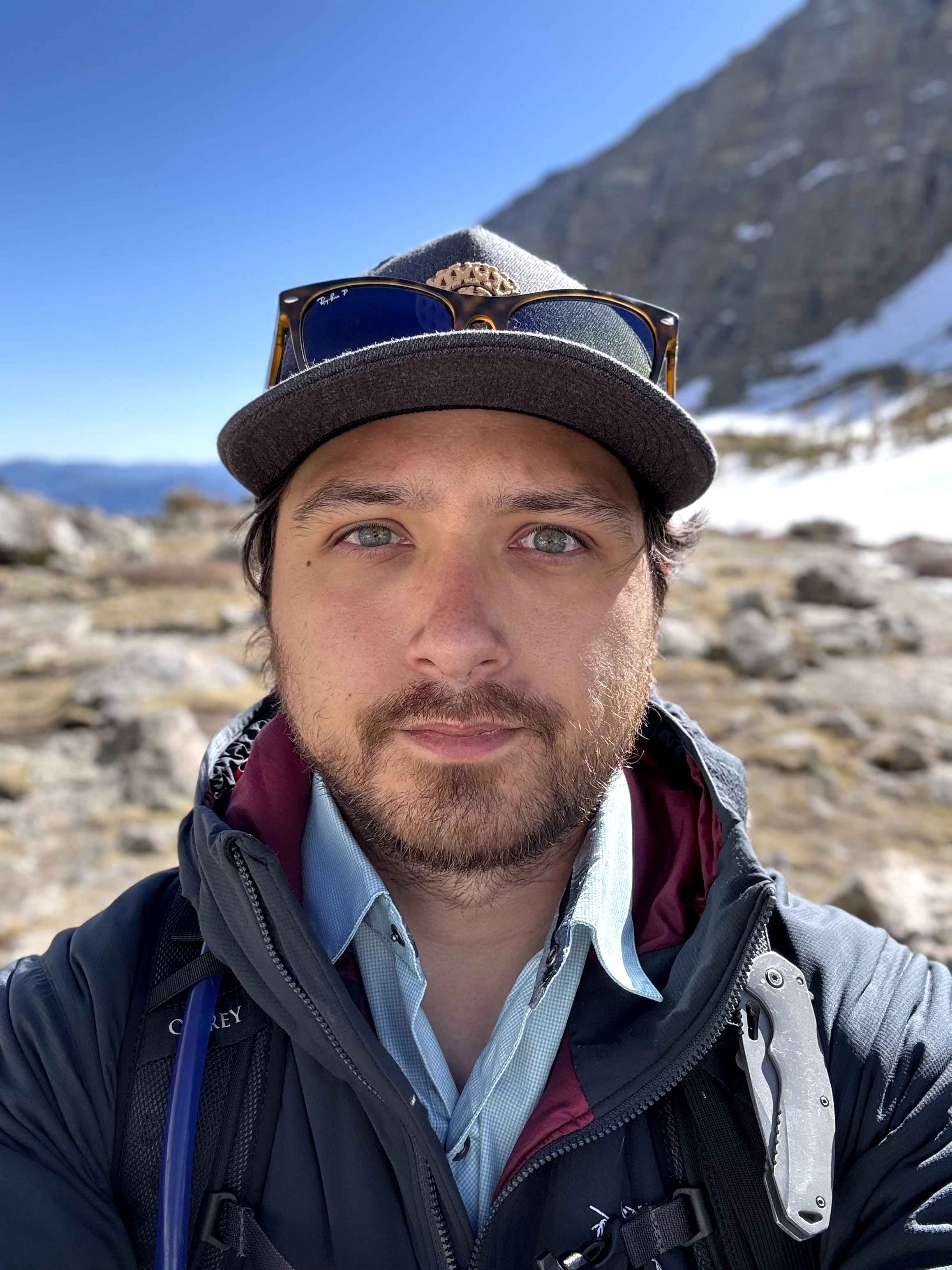 Outdoor selfie of a person wearing a cap, hiking jacket, and backpack, standing in a rocky mountain landscape with snow patches and a clear blue sky.