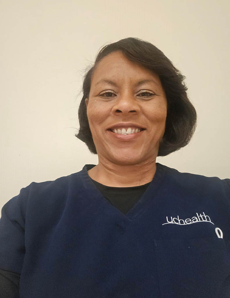 Smiling person in navy UCHealth scrubs over a black shirt, with short dark hair, facing the camera against a plain light-colored wall in a well-lit indoor setting.
