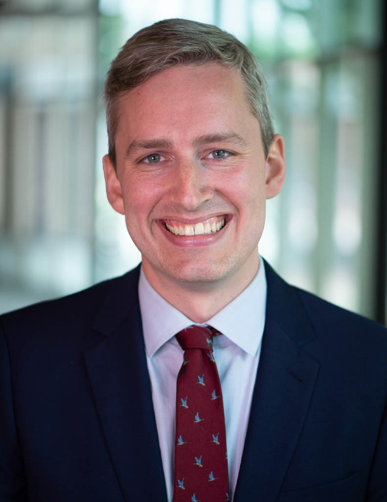 Headshot of a smiling man with short light brown hair wearing a navy suit, light blue shirt, and red patterned tie, standing in front of a softly blurred indoor background.