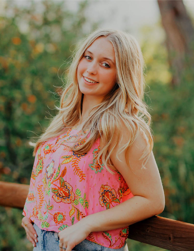 Outdoor portrait of a smiling woman with long blonde hair wearing a pink floral sleeveless top and jeans, leaning against a wooden railing with greenery in the background.