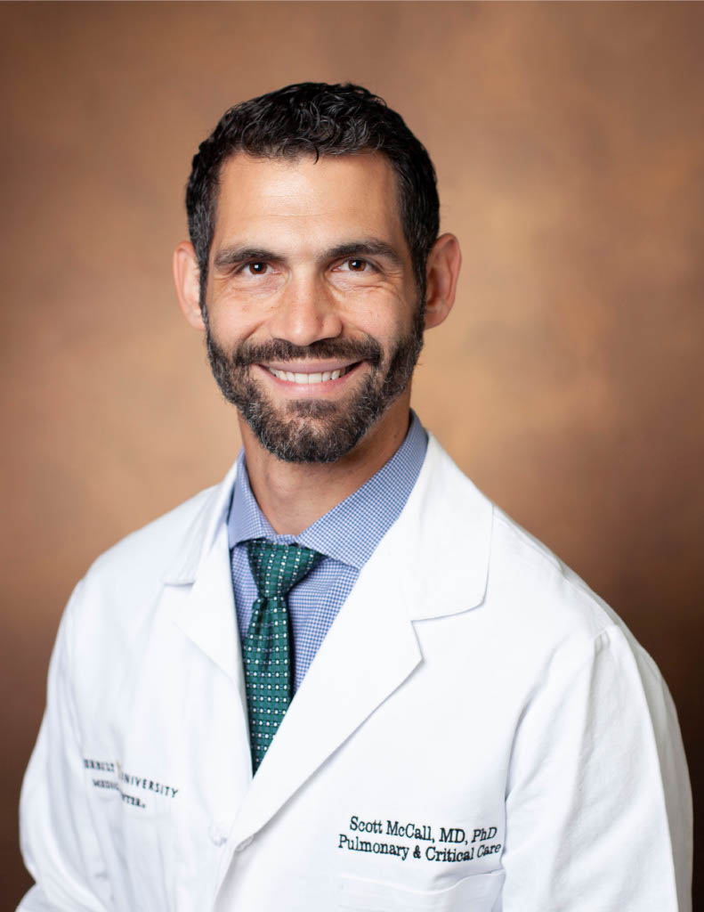Professional headshot of a smiling physician with short dark hair and a beard wearing a white lab coat, dress shirt, and green tie against a brown studio background.