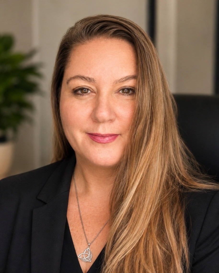 Professional headshot of a woman in a black blazer with long hair, smiling in an office setting.