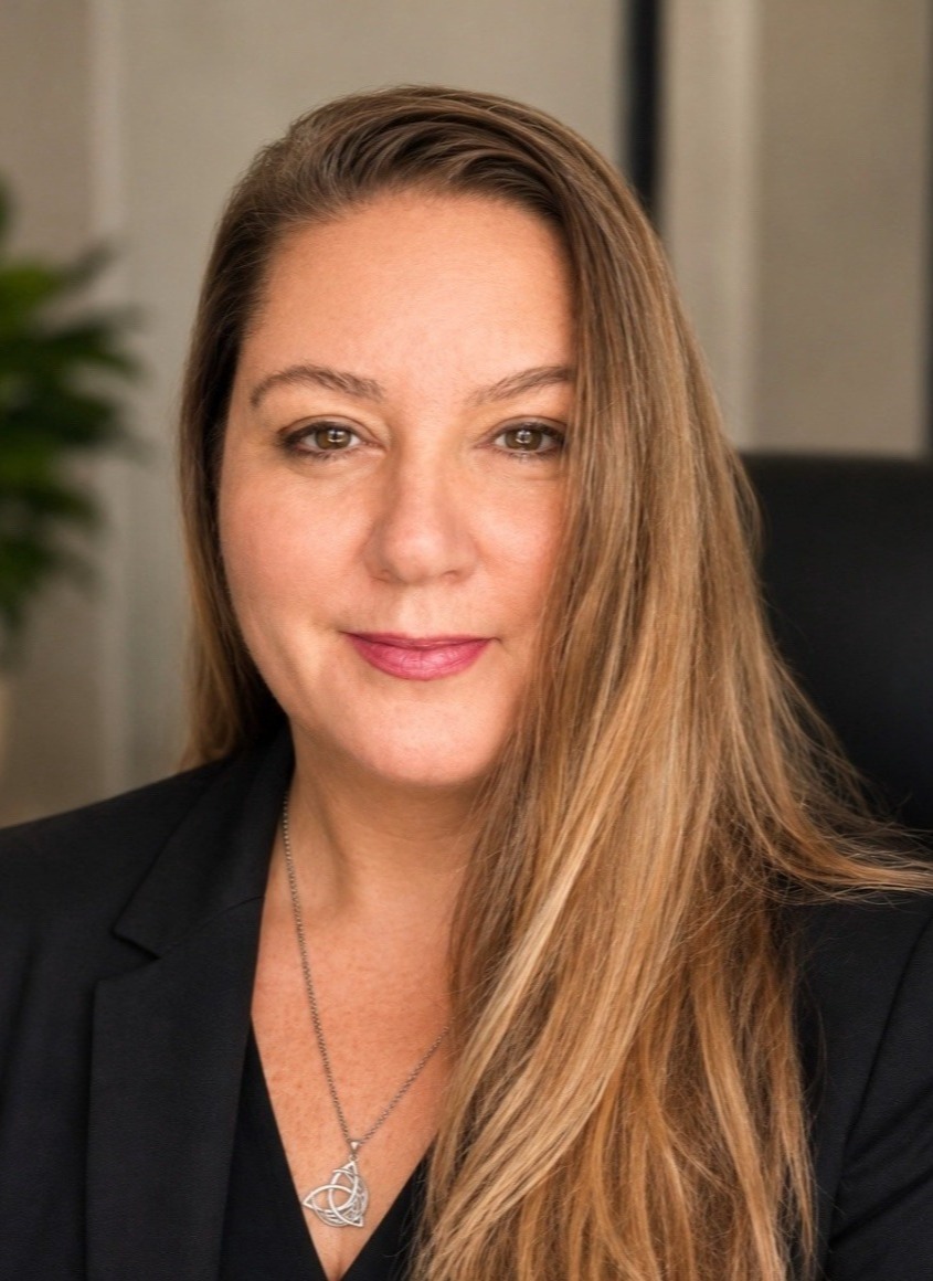 Professional headshot of a woman in a black blazer with long hair, smiling in an office setting.