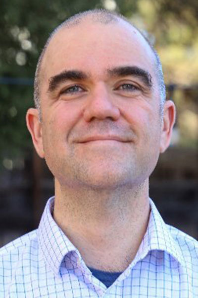Professional headshot of a smiling man wearing a light checkered button-down shirt, photographed outdoors with a blurred natural background.