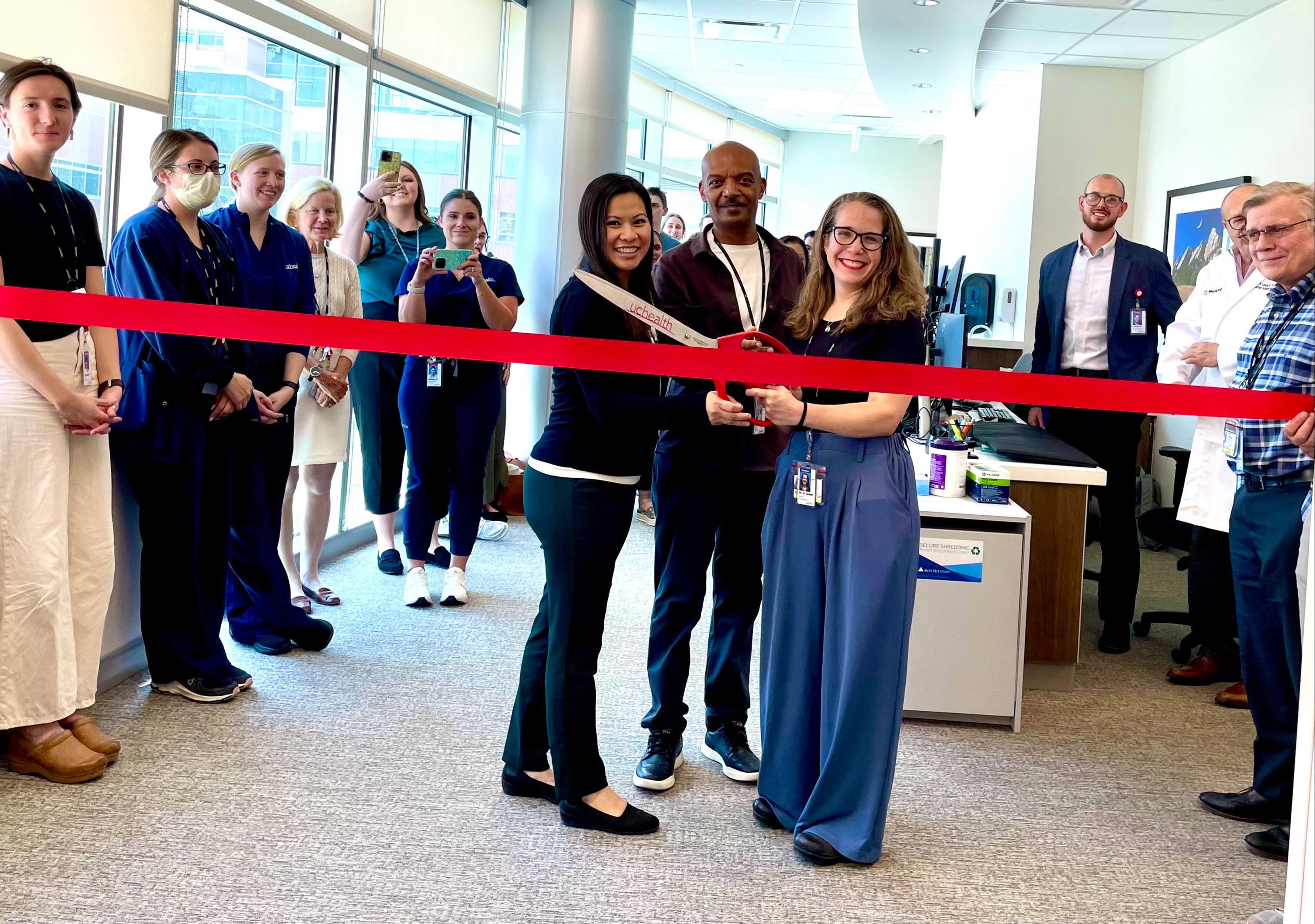 Photo of several people at a clinic opening. 3 individuals are cutting a red ribbon in front.