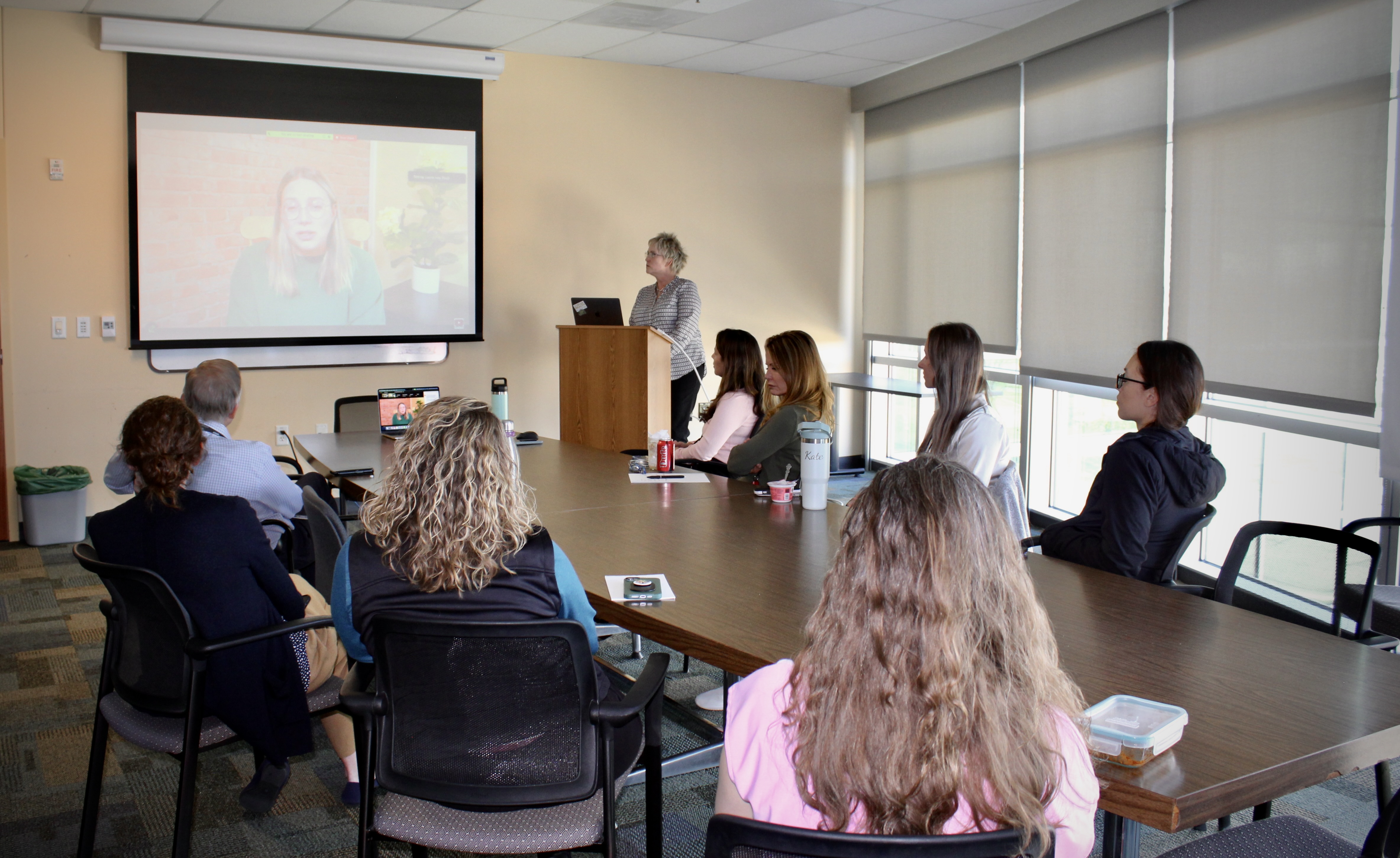 Photo of 9 individuals in a conference room. 8 Sitting at a desk, 1 at podium. There is a projector screen with a zoom participant.