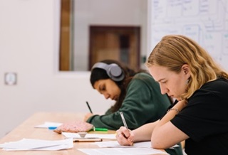 Anxiety - Photo by Jeswin Thomas on Unsplash Two students sitting at a table writing. One student has earphones.