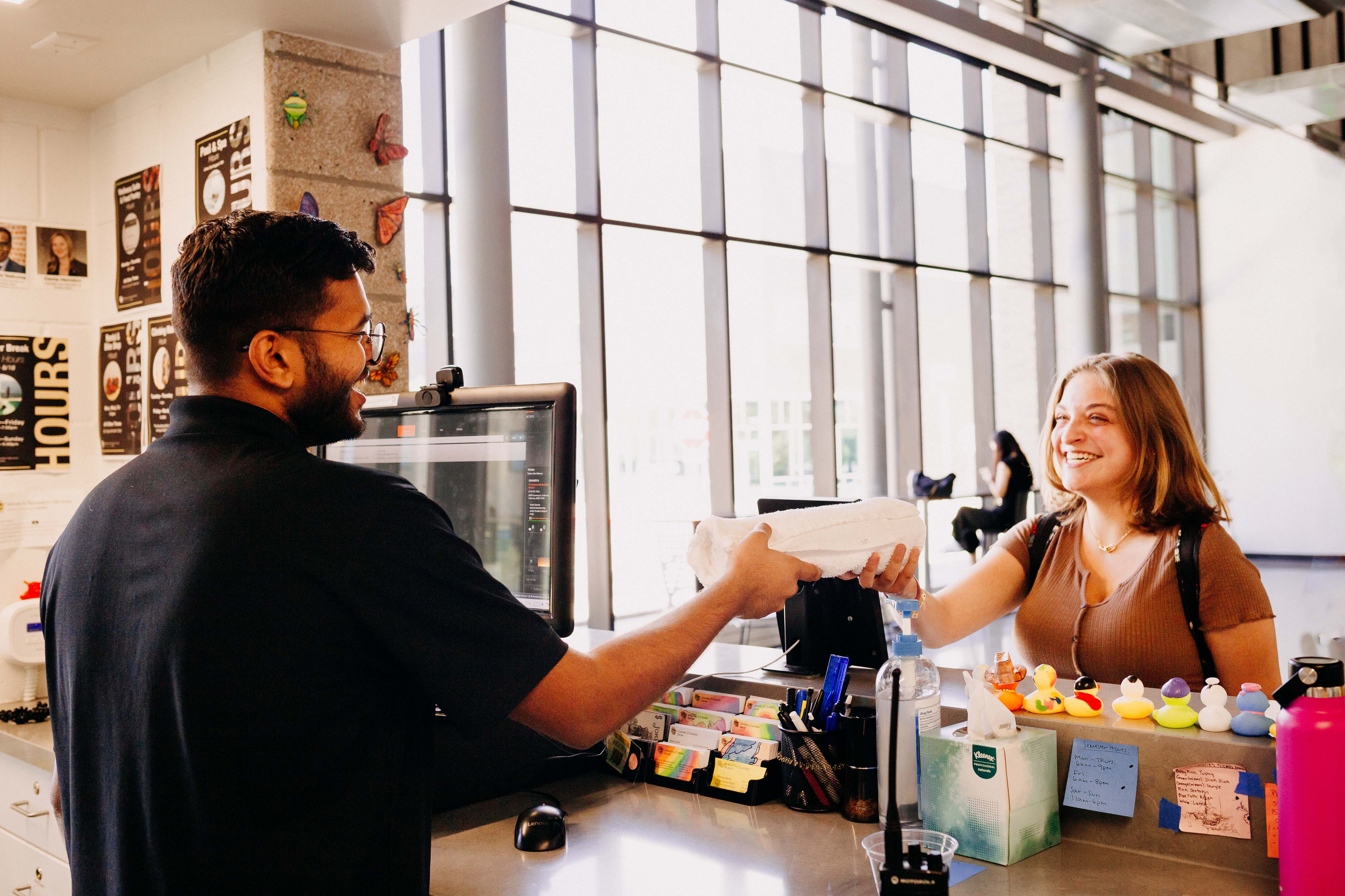 A smiling staff member behind a reception desk handing a white towel to a happy student.
